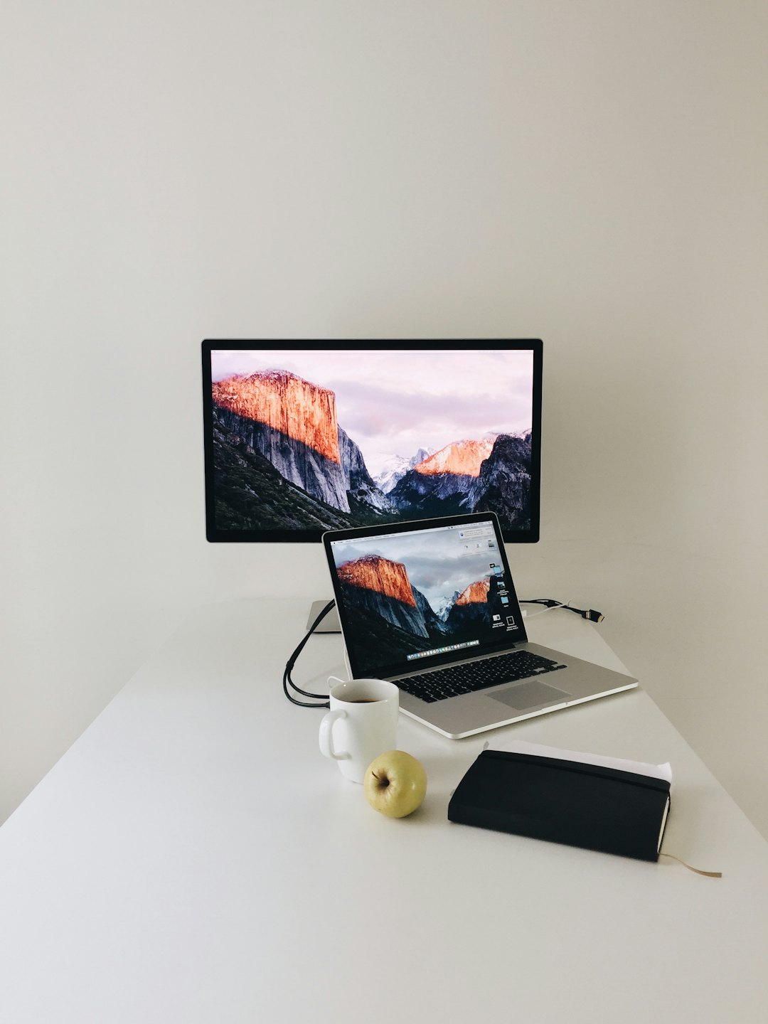 Minimalistyczne biurko z laptopem, monitorem, jabłkiem i kubkiem kawy.